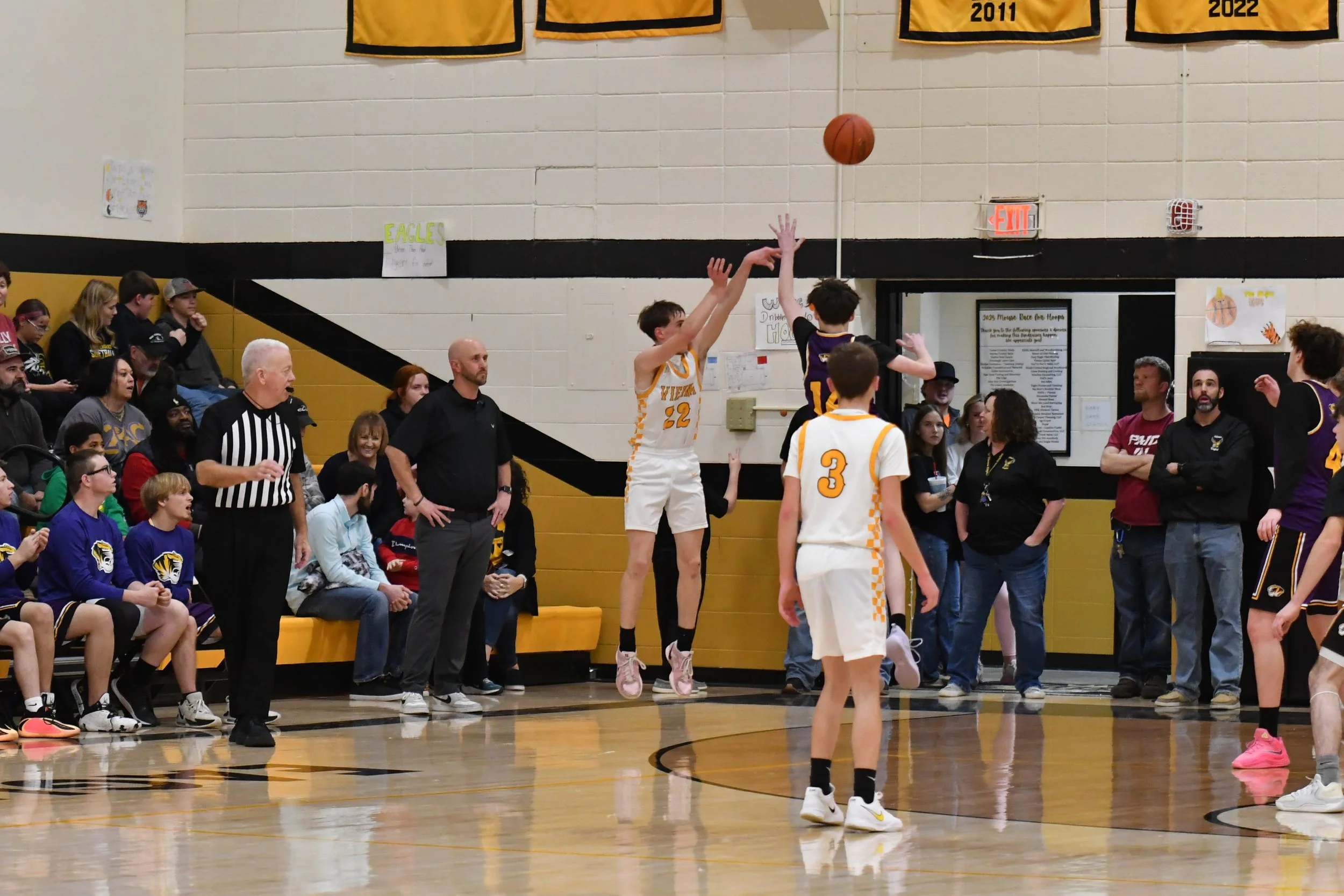 A basketball game with players in black and white uniforms, one player in mid-air taking a shot, and others watching on the court. Spectators and coaches are seated and standing along the sidelines, with banners hanging on the wall behind them.