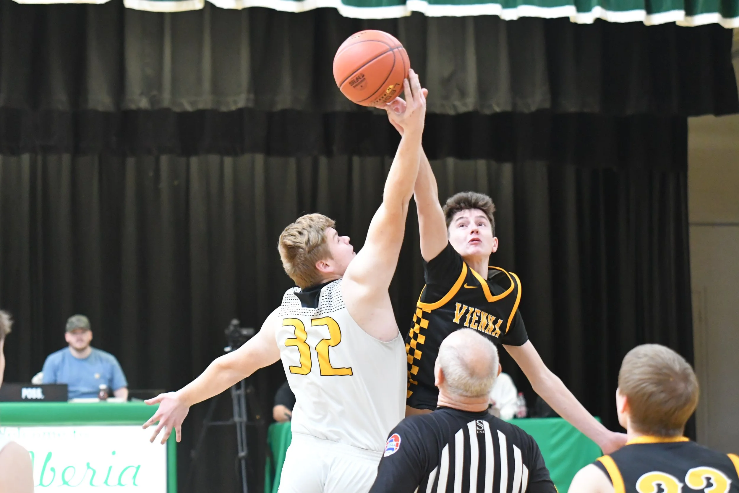 Two basketball players, one in a white jersey and the other in a black jersey with yellow accents, jump for the ball at the tip-off, with a referee in a black and white striped shirt observing. The white jersey player has the number 32, and the black