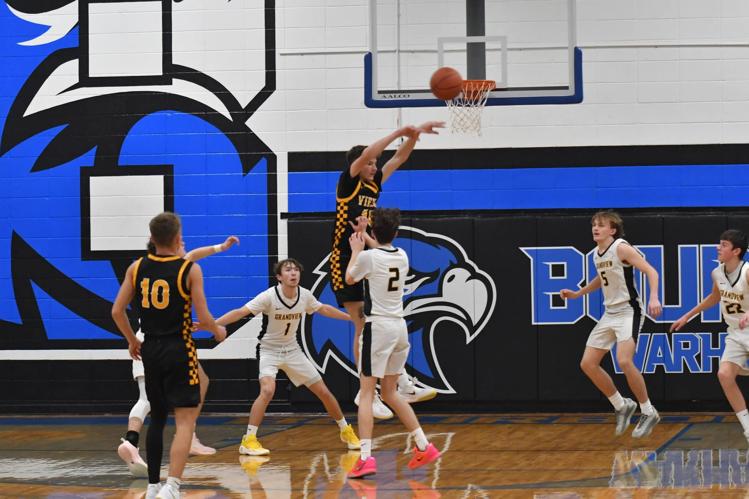 Kids playing basketball in a gymnasium, with one player shooting the ball near the basket while others are defending or watching.
