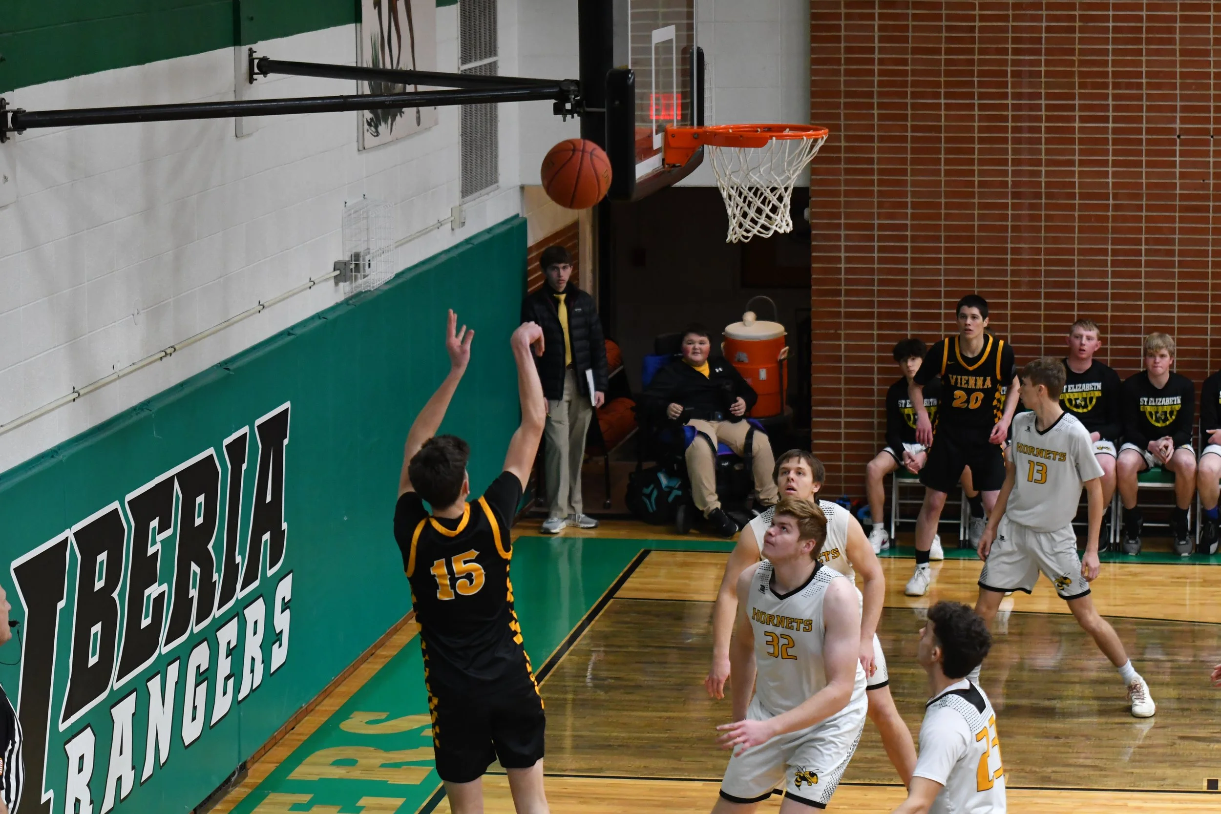 A basketball player in black jersey number 15 shoots a basketball towards the hoop as players and spectators watch during a game in a gymnasium.