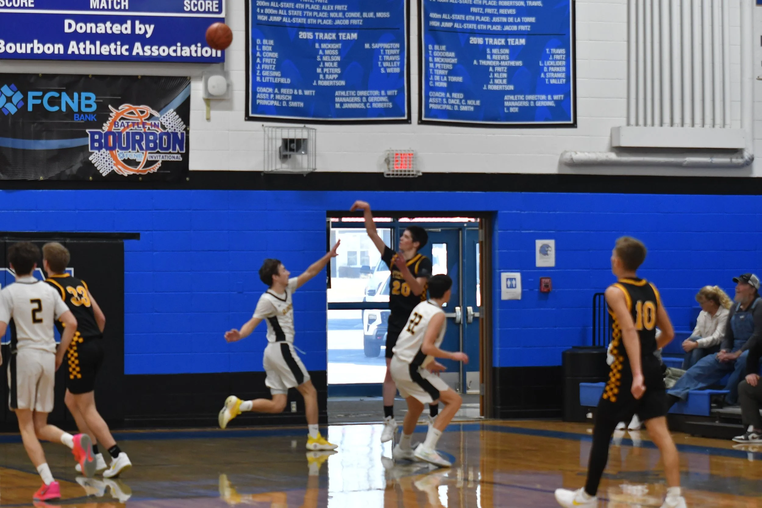 A boys' basketball game in progress inside a gymnasium. Players are actively participating, with one attempting a shot while others are defending or moving on the court. The gym has blue walls with banners and posters, including blue scoreboard banne