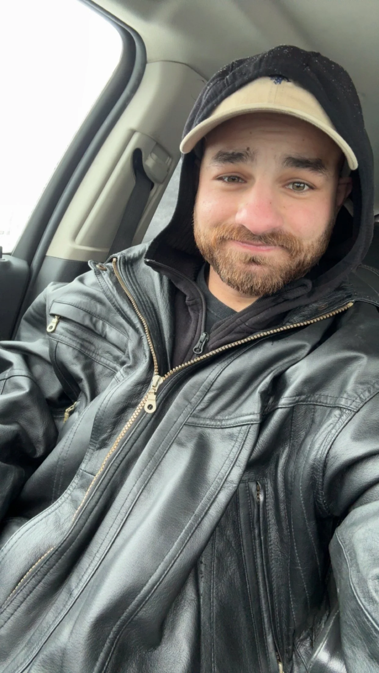 A young man with a beard, smiling, wearing a black hoodie, a beige cap, and a black leather jacket, sitting inside a vehicle.