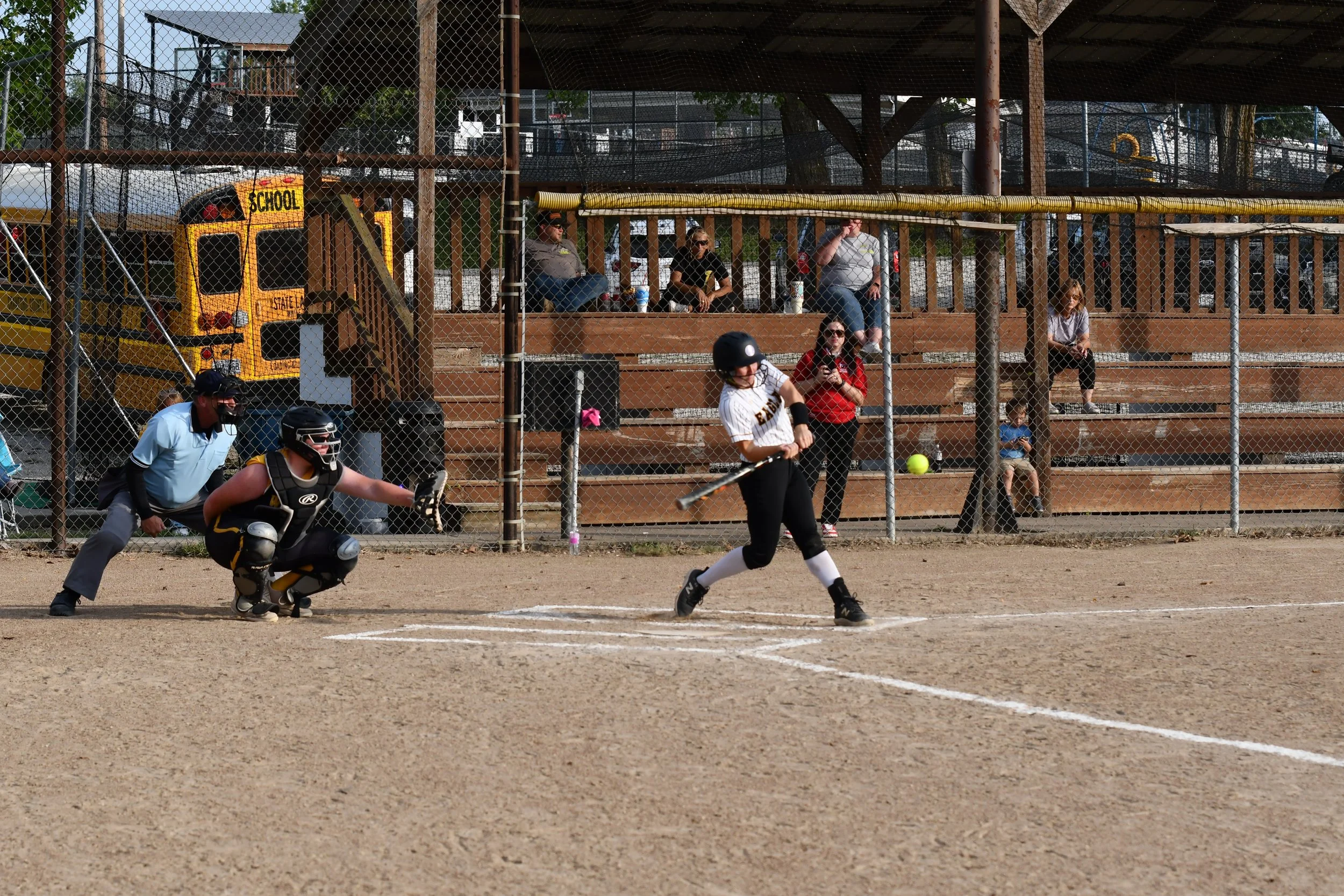 A girl batting in a softball game, with a catcher and umpire behind her. Spectators are sitting on bleachers in the background, and a school bus is parked behind the fence.