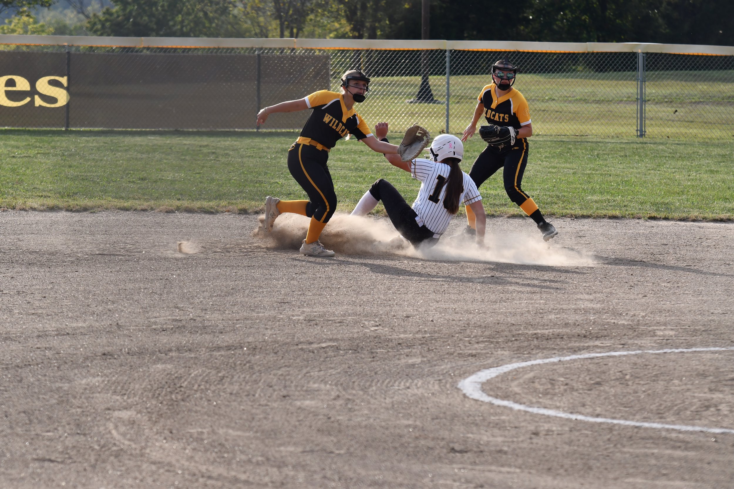 Softball players in black and yellow uniforms attempt to tag a runner in a white and black uniform sliding into a base, with two other players nearby on the field.