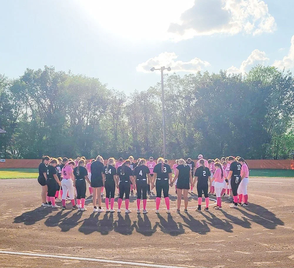 Softball players in uniforms holding hands in a circle on a dirt field during daylight.