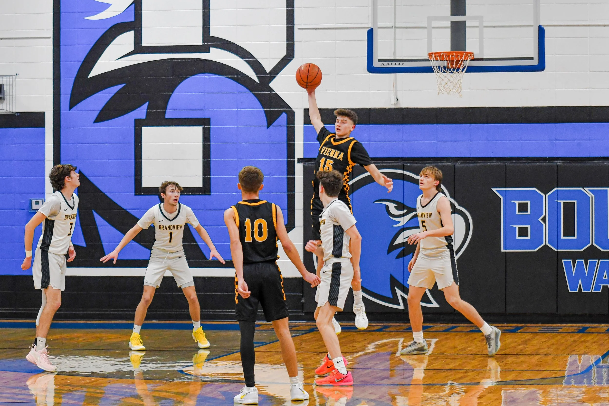 A group of young boys playing basketball in a gymnasium. One player in a black and yellow uniform jumps to make a shot, while others in white uniforms try to block or defend. The gym has a blue and black mascot/logo on the wall.