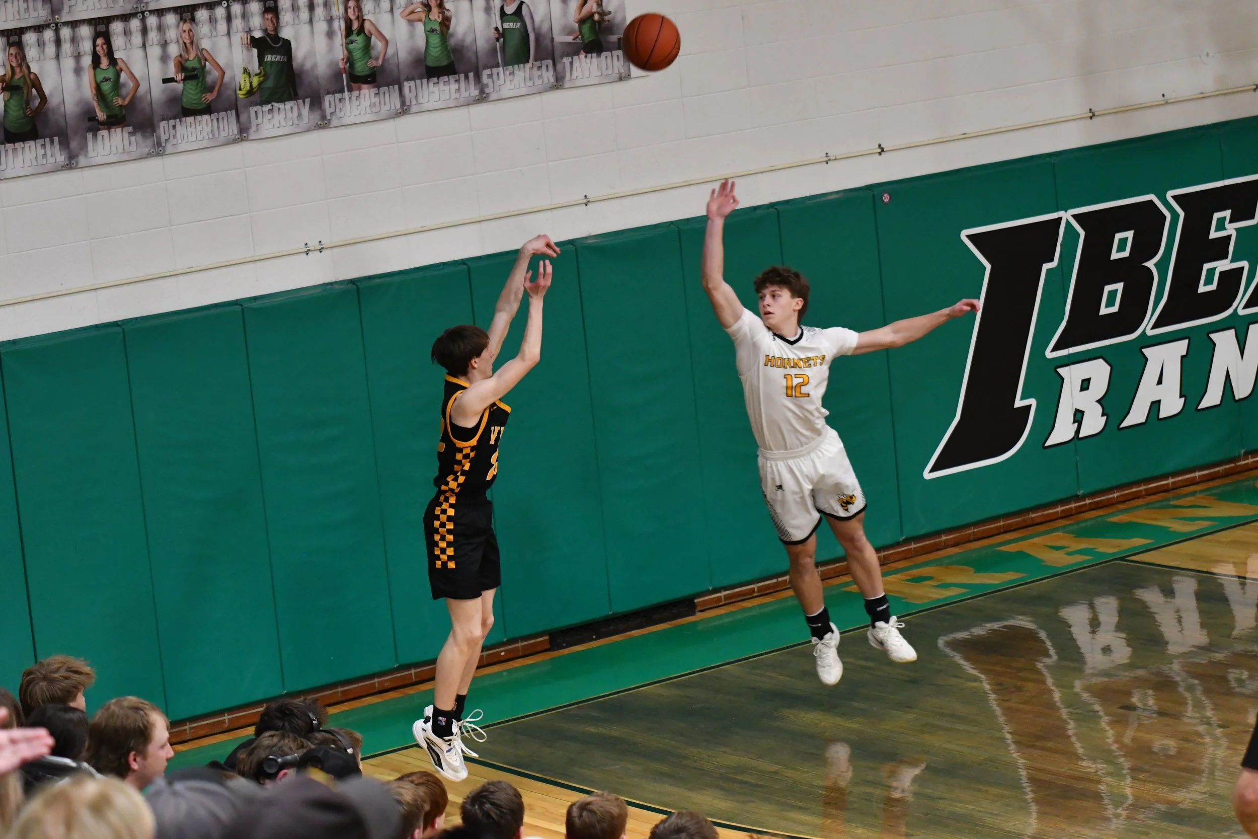Two basketball players are jumping in the air for a rebound during a game in a gymnasium. One player is wearing a black and yellow uniform, while the other is wearing a white uniform with yellow and black accents. Spectators are sitting on the floor 
