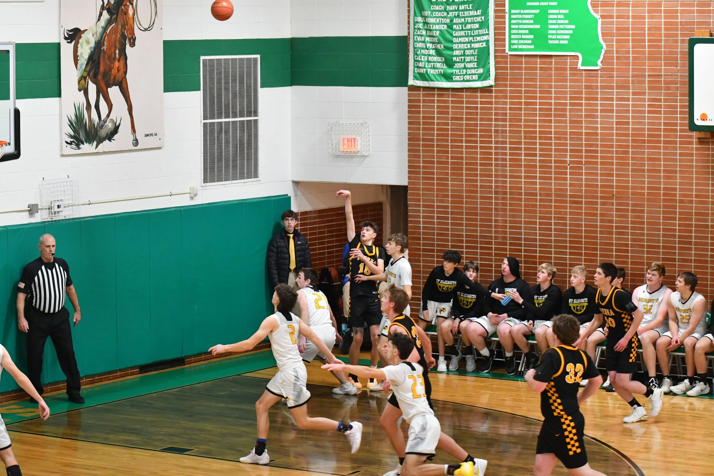 Young basketball players in a game, with some attempting to catch a ball near the wall and others on the bench watching, inside a gymnasium with brick and panel walls, a mural of a horse, and green banners hanging.