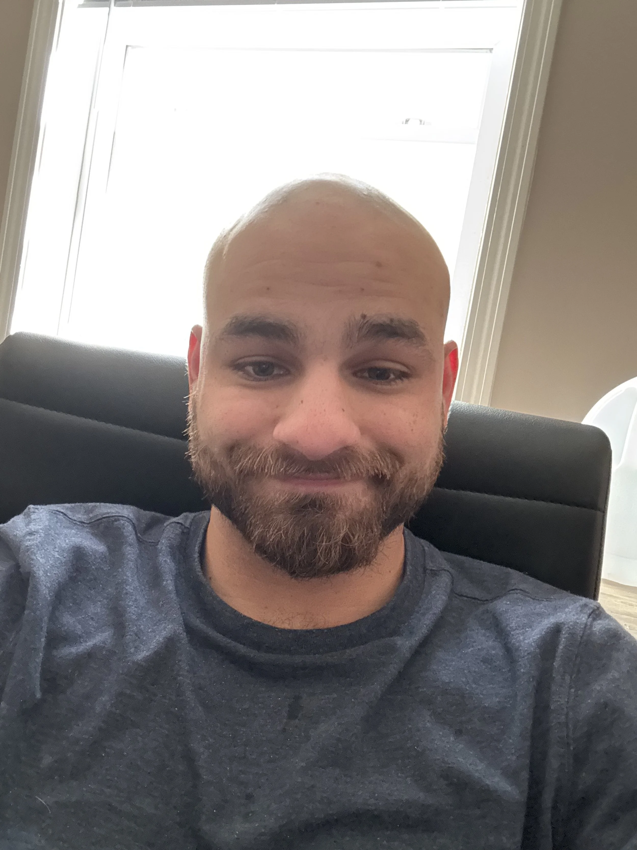 A young man with a beard and shaved head sitting on a black chair, smiling slightly. Behind him, a window with light shining through and part of a white object is visible.