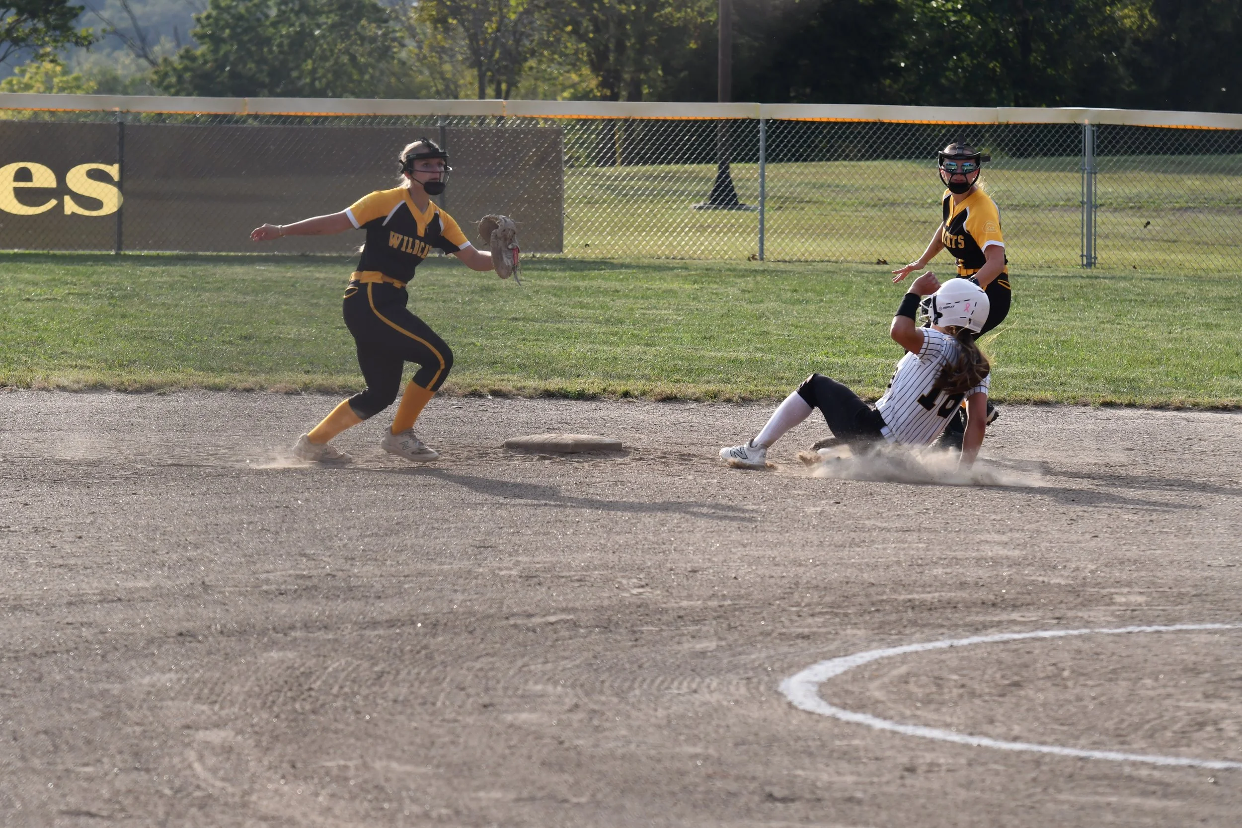 A softball game with players in black and yellow uniforms, one sliding into a base, another preparing to catch, and a third approaching, all on a dirt field with green grass and trees in the background.