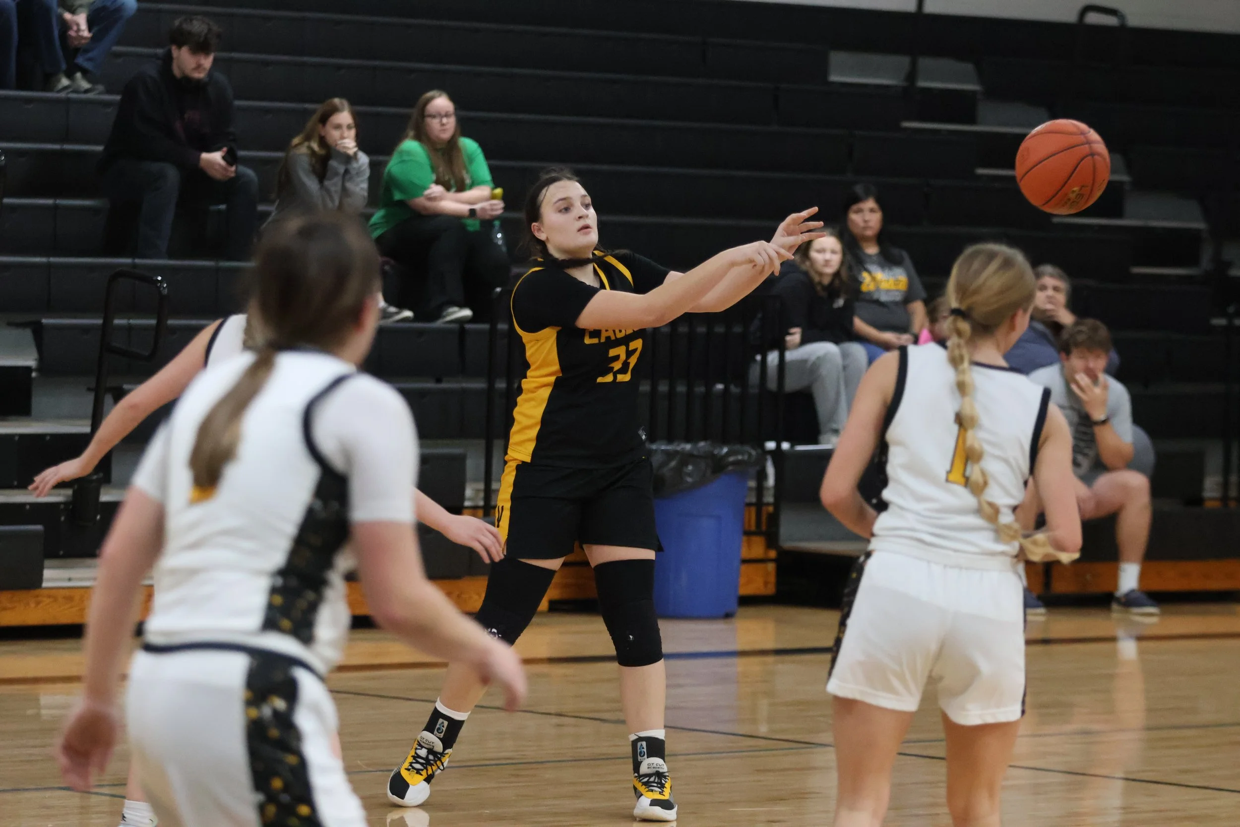 A women's basketball game in progress showing players and spectators. One player in a black and yellow uniform is passing the ball, while two players in white uniforms with black and yellow accents are on court. Spectators are sitting on black bleach