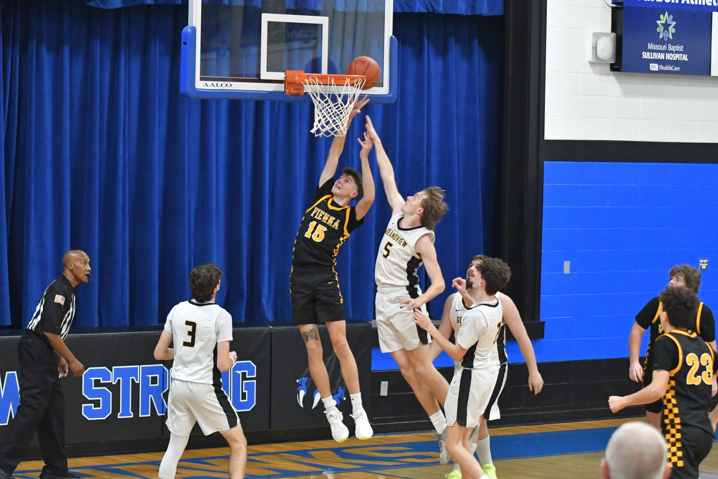 A basketball game where players are jumping near the hoop. One player in black jersey number 16 is attempting a shot while a player in white jersey number 5 is trying to block it. Other players are watching and preparing for the play. An official ref