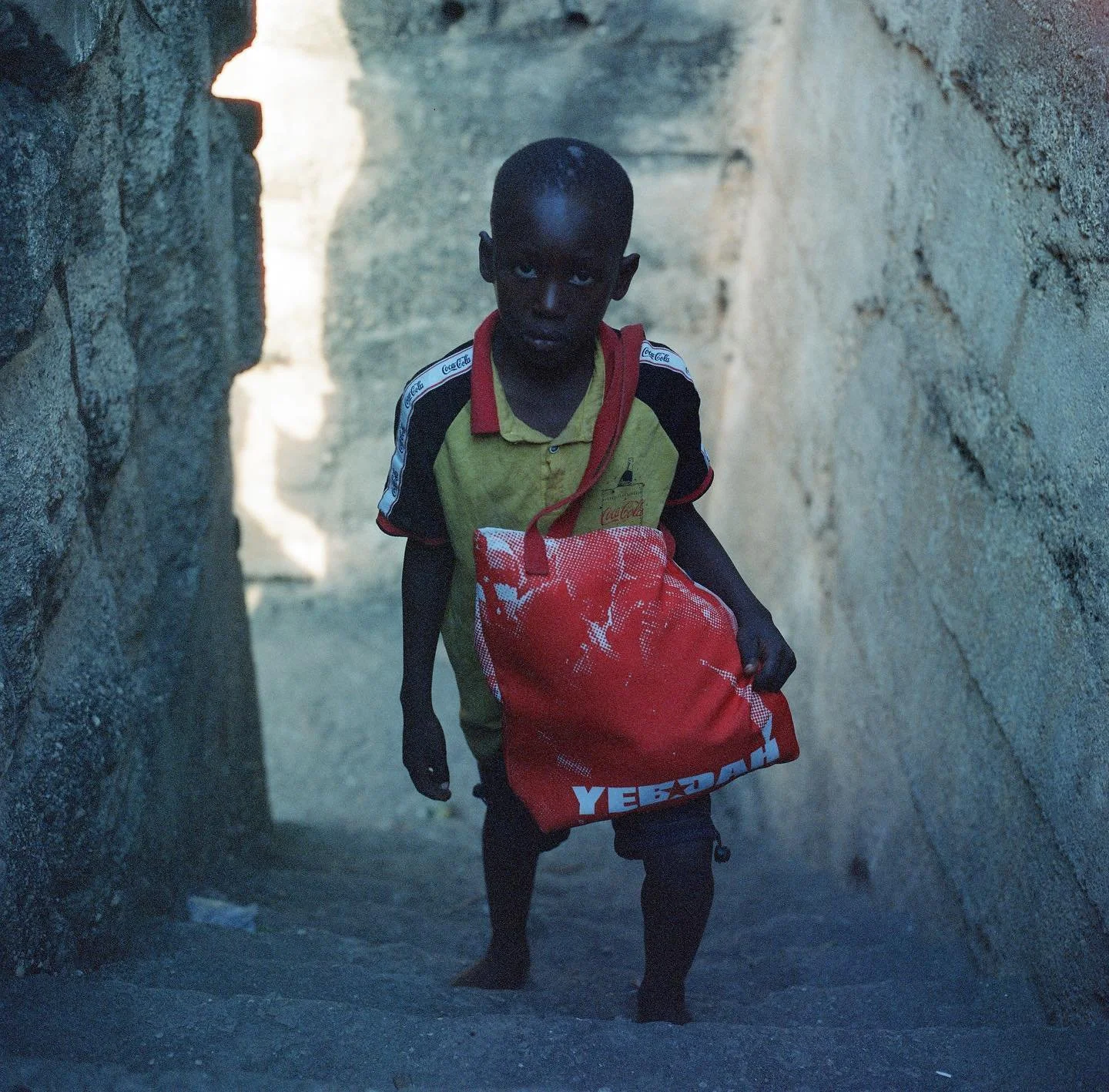 A young boy walking through a narrow stairway between stone walls, carrying a red bag with white lettering reading YEBOAH Studios and wearing a yellow and black shirt.