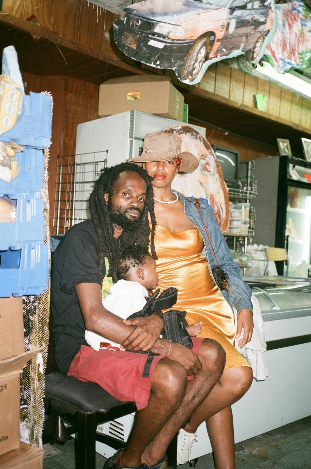 A family inside an African Market in NYC, with a man holding a child on his lap and a woman leaning beside them. The man has dreadlocks and a beard, the woman wears a large hat and an orange dress, and the child is leaning against the man.