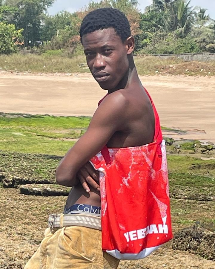 A young man standing outdoors in a field with green grass and rocks, wearing beige pants, a visible Calvin Klein waistband, and a red YEBOAH tote bag over his shoulder.