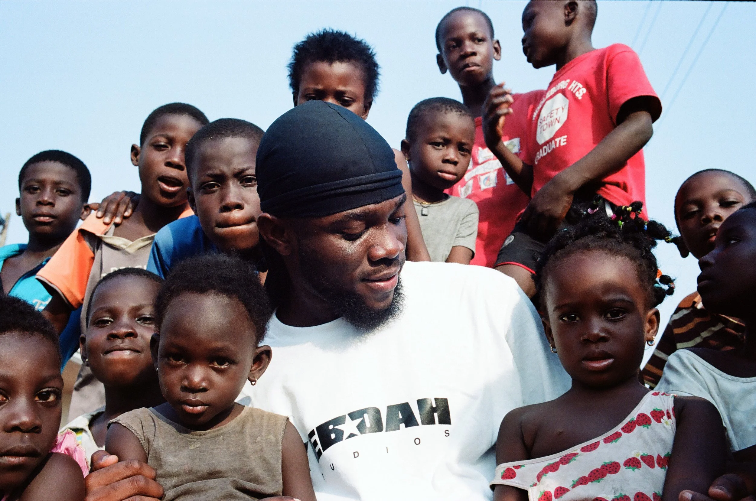 A man surrounded by several young children with a clear blue sky in the background, with some children gently touching his shoulders and head, and looking at the camera and at the man.