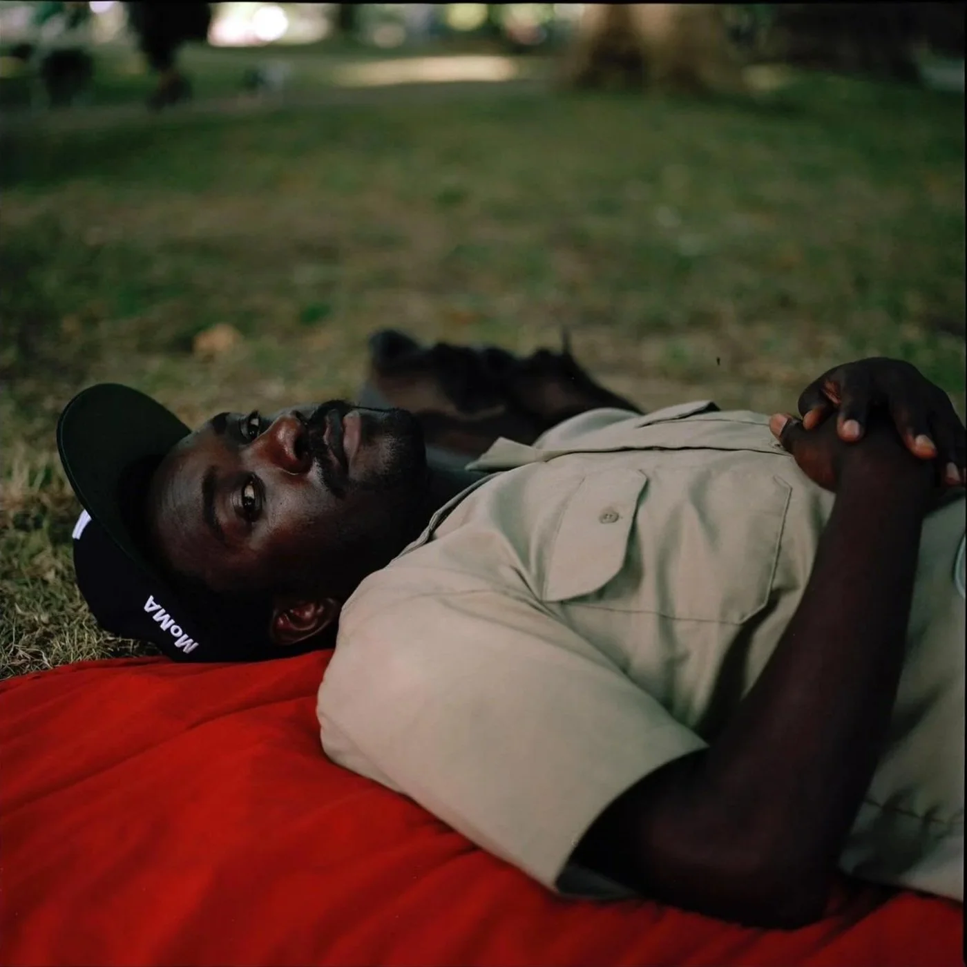 A man, Lloyd Foster, lying on the ground with hands clasped, looking upward in a grassy outdoor setting, wearing a khaki shirt and a cap.