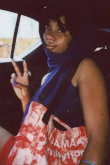 A young woman sitting in a car, making a peace sign with her right hand, wearing a dark blue scarf and a red and white patterned bag with the word 'YEBOAH' visible.
