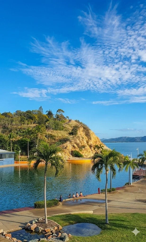 Scenic view of a waterfront with palm trees, a rocky hillside, and a clear blue sky with scattered clouds.