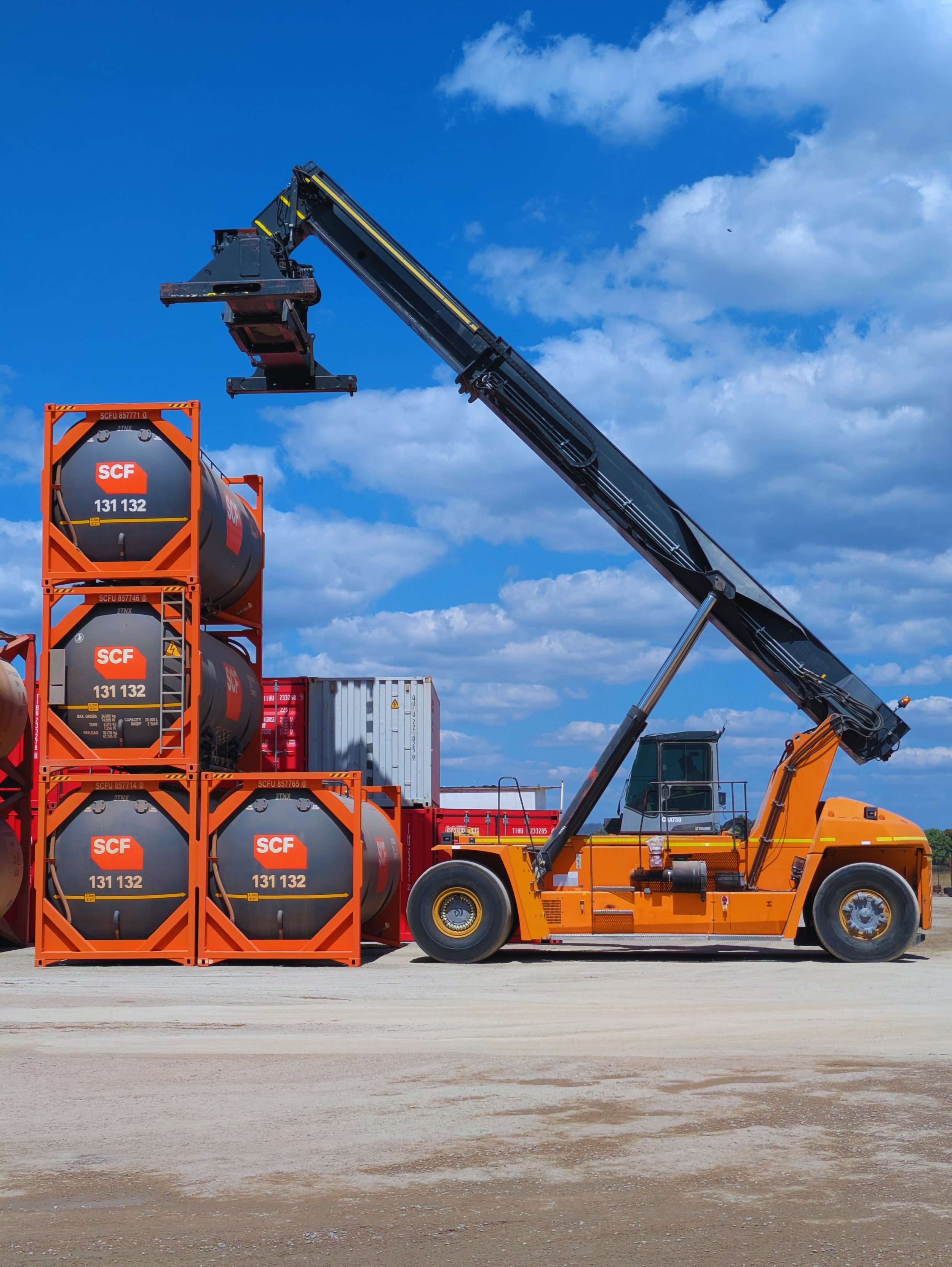A construction scene with an orange truck lifting large black tanks labeled 'SCF 131 132' onto orange storage racks. There are additional containers and equipment in the background under a blue sky with scattered clouds.