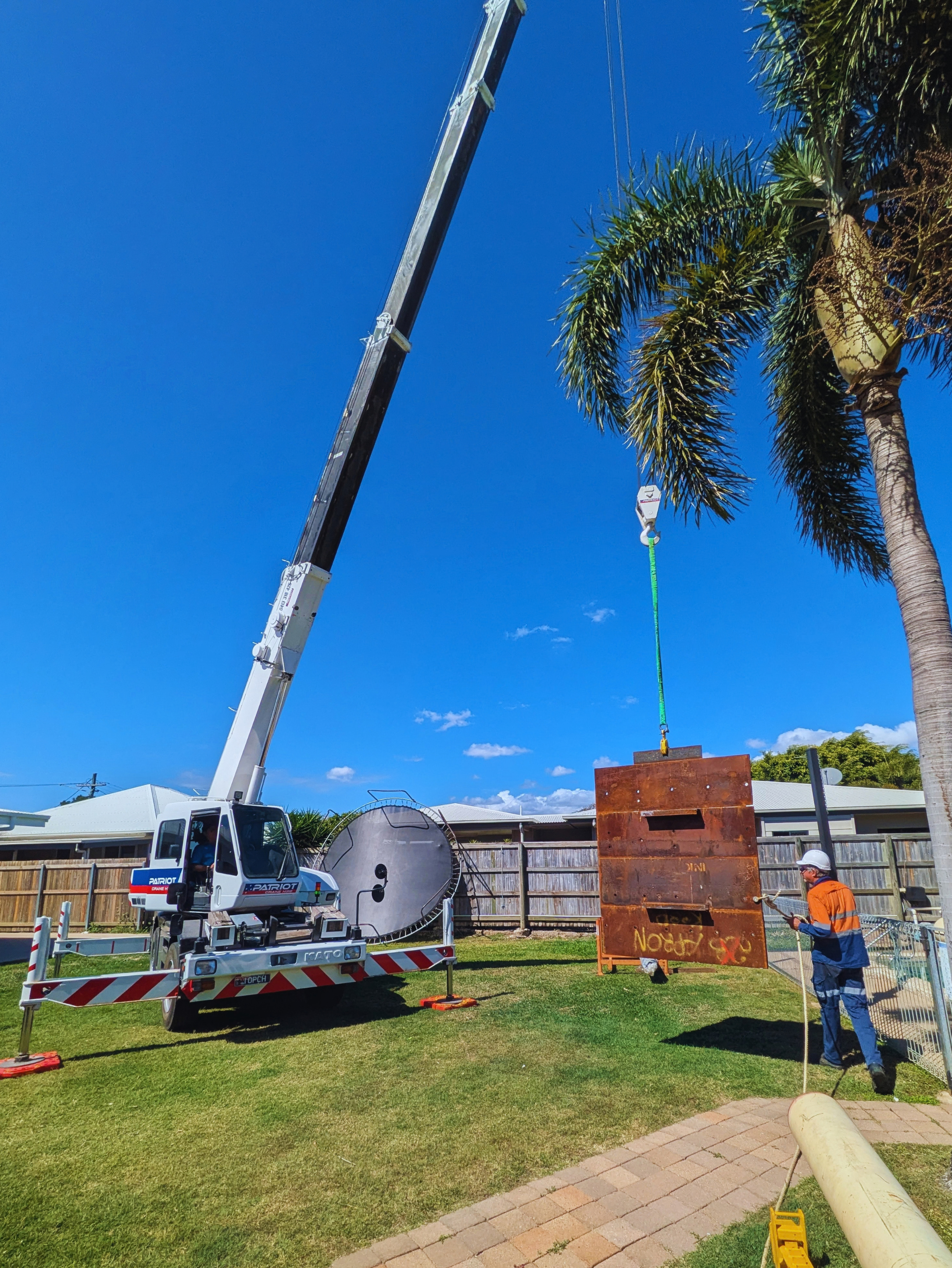 A crane truck outdoors lifting a large rusted metal structure with workers nearby, lush green grass, a wooden fence, and a house with a white roof in the background.