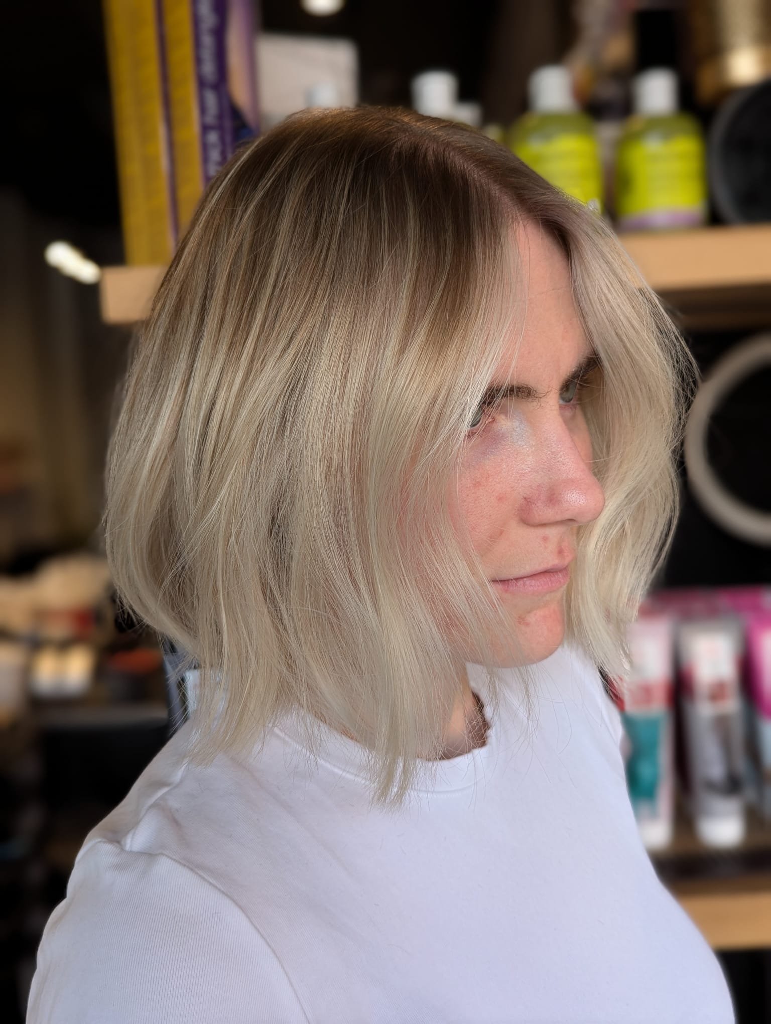 A woman with shoulder-length blonde hair with darker roots, wearing a white shirt, standing in a store with hair products on shelves in the background.