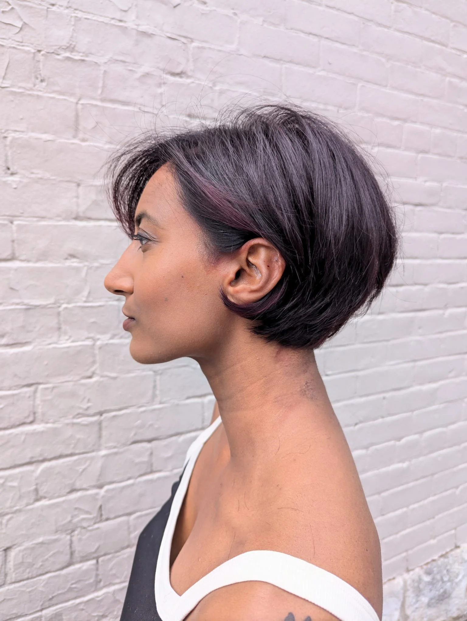 Side profile of a woman with short, dark purple hair with pink highlights, standing against a white brick wall.