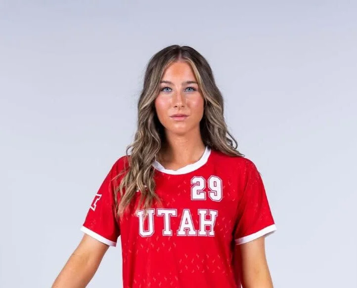 A young woman with long wavy blonde hair is wearing a red sports jersey with white accents, the word 'UTAH' and the number 29. She has a neutral expression and is against a plain light gray background.