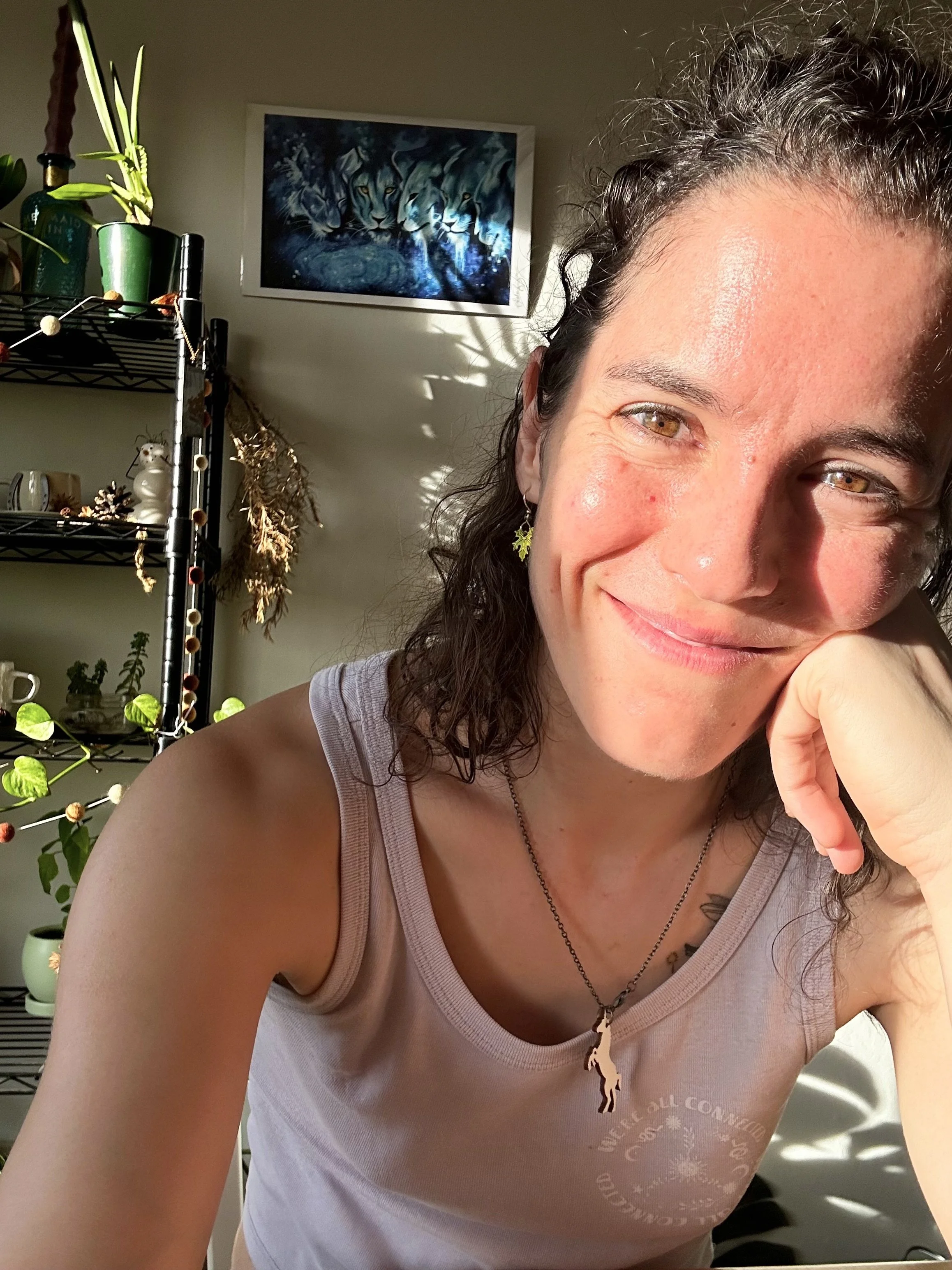 Caitlin Clausen smiling softly, chin resting on her hand, in warm golden light with plants and dried flowers behind her.