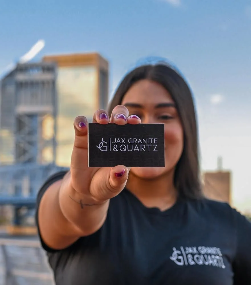 Mujer sosteniendo una tarjeta con el logo de Jax Granite & Quartz frente a la cámara, fondo urbano con cielo azul.