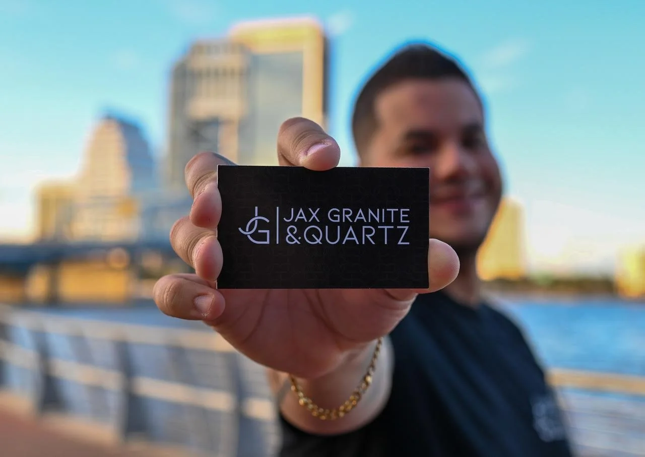 Hombre sosteniendo una tarjeta que dice 'JAX GRANITE & QUARTZ' con río y edificio de ciudad de fondo