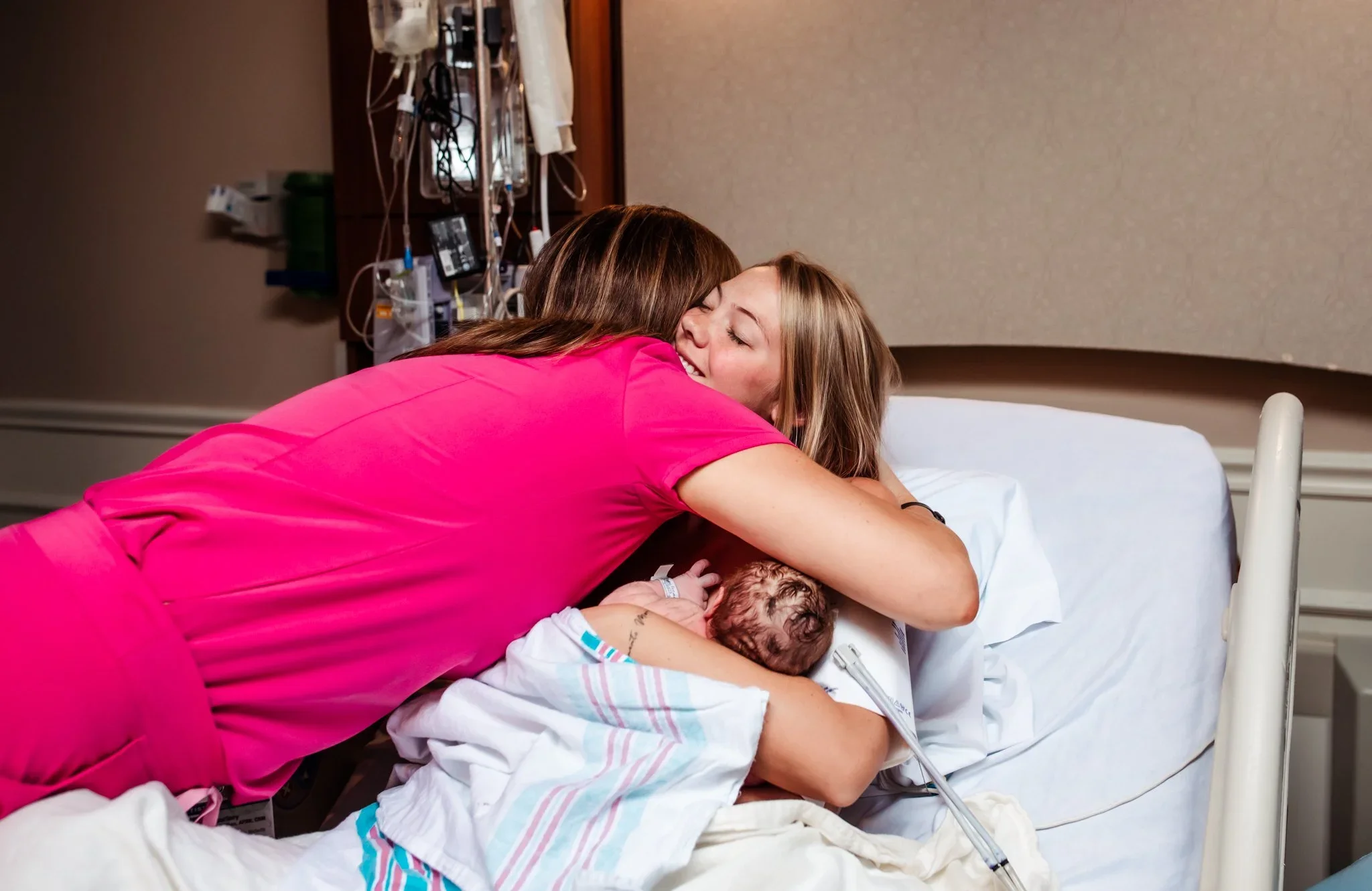 A woman in pink scrubs is hugging a woman in a hospital bed after childbirth, who is holding a newborn baby.