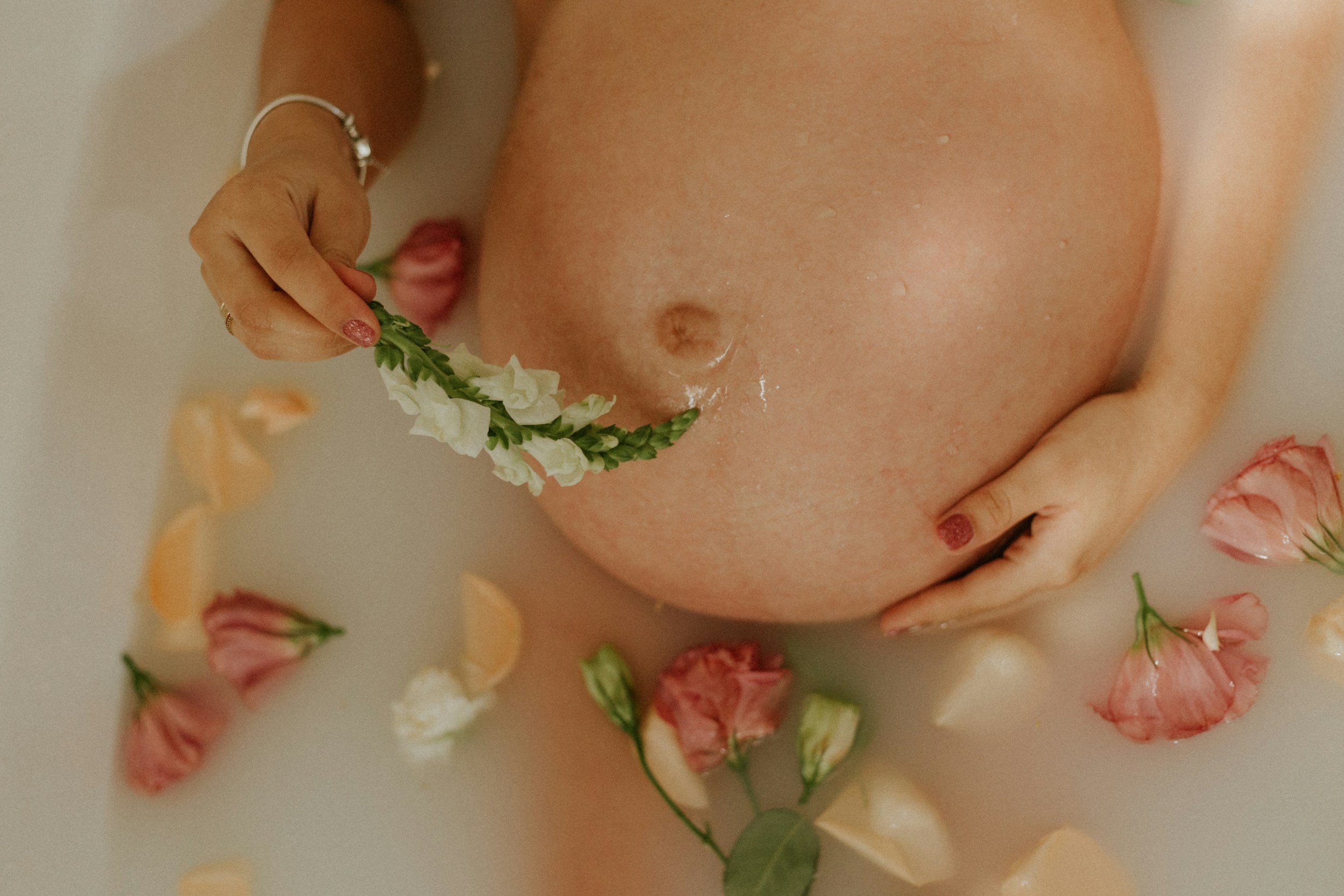 A pregnant woman is relaxing in a milk bath surrounded by pink and white flowers and petals, holding a flower near her belly.
