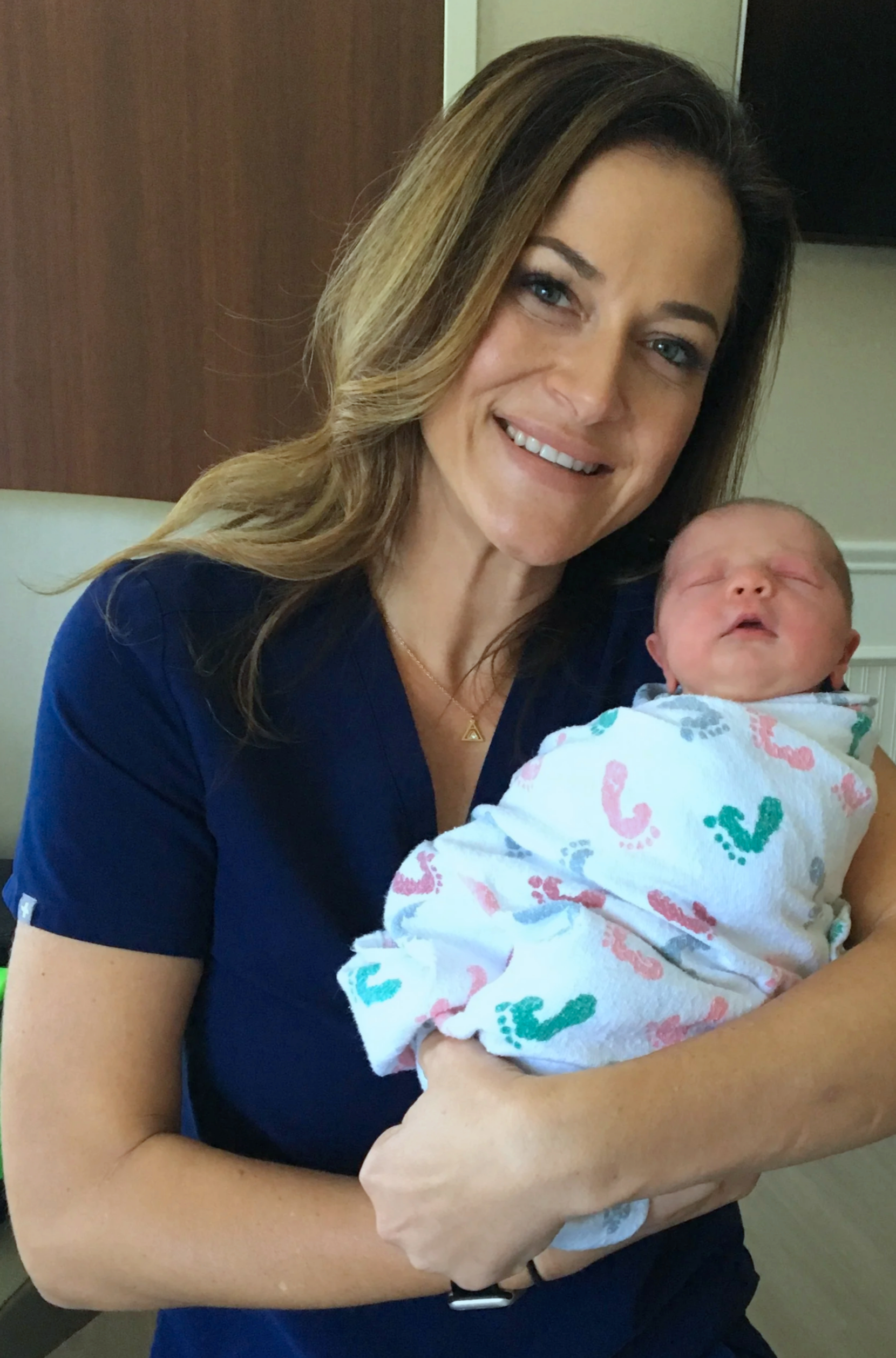 Midwife holding a newborn baby wrapped in a hospital blanket, smiling at the camera.