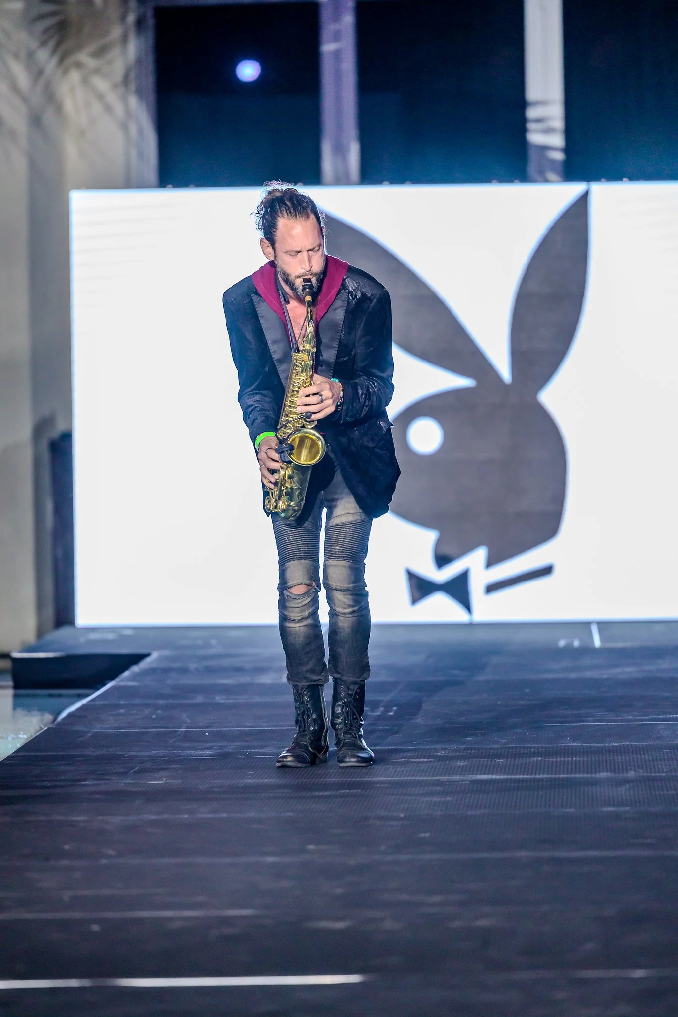 A man walking on a runway playing a saxophone, with a large black and white PlayBoy bunny logo in the background.