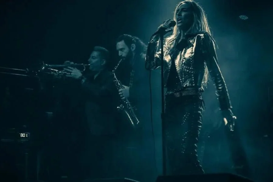 A live performance with a female singer in a leather outfit singing into a microphone, accompanied by two male musicians playing trumpet and saxophone on stage with blue lighting.