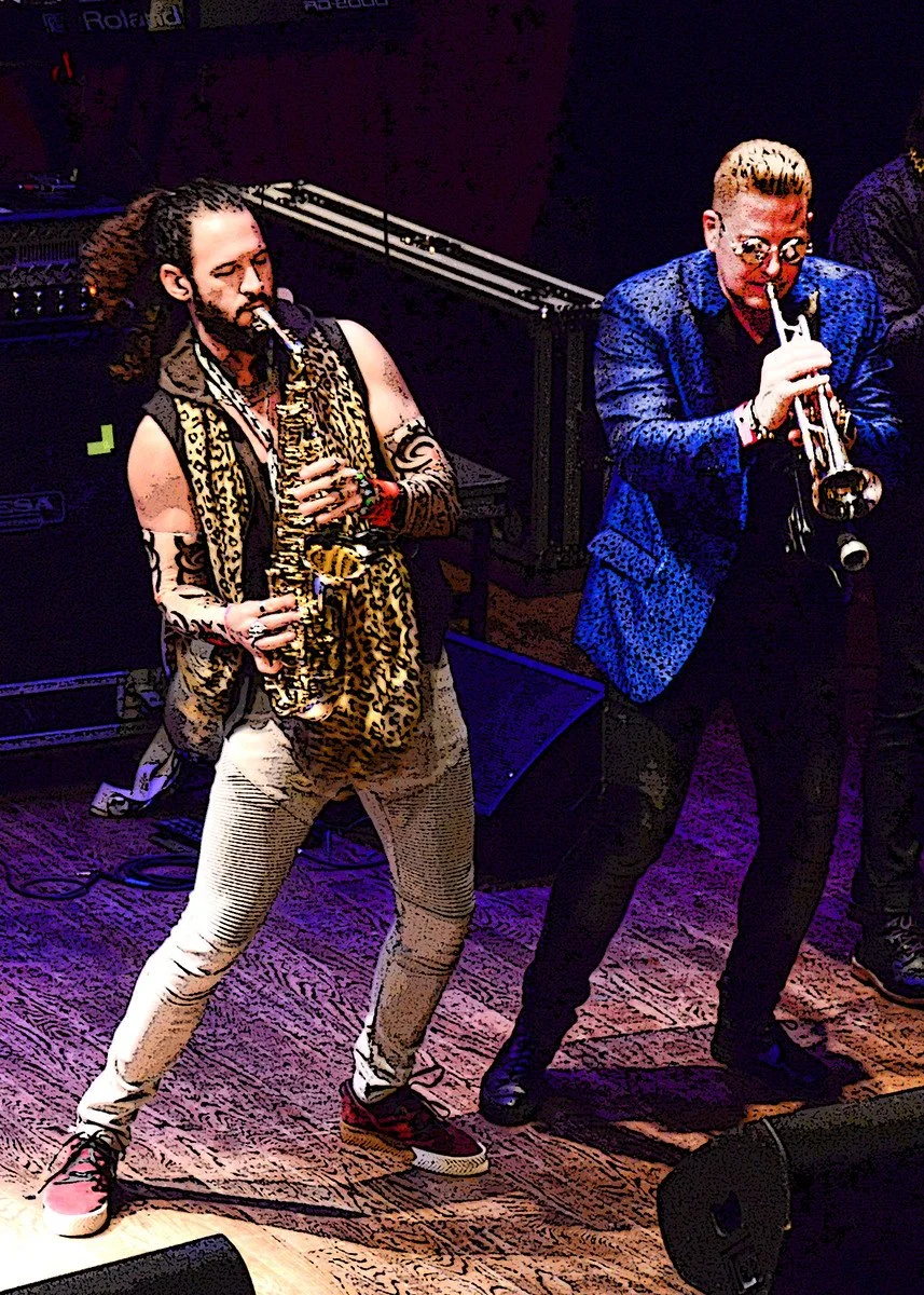 Two musicians playing wind instruments on stage, one with a saxophone and the other with a trumpet, amid stage equipment and colorful lighting.