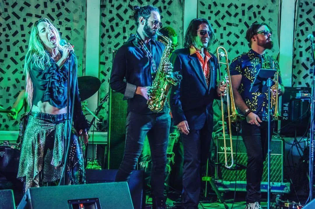 A band performing live on stage with female lead singer, saxophonist, trombonist, and another musician playing a keyboard, all under colorful stage lighting.