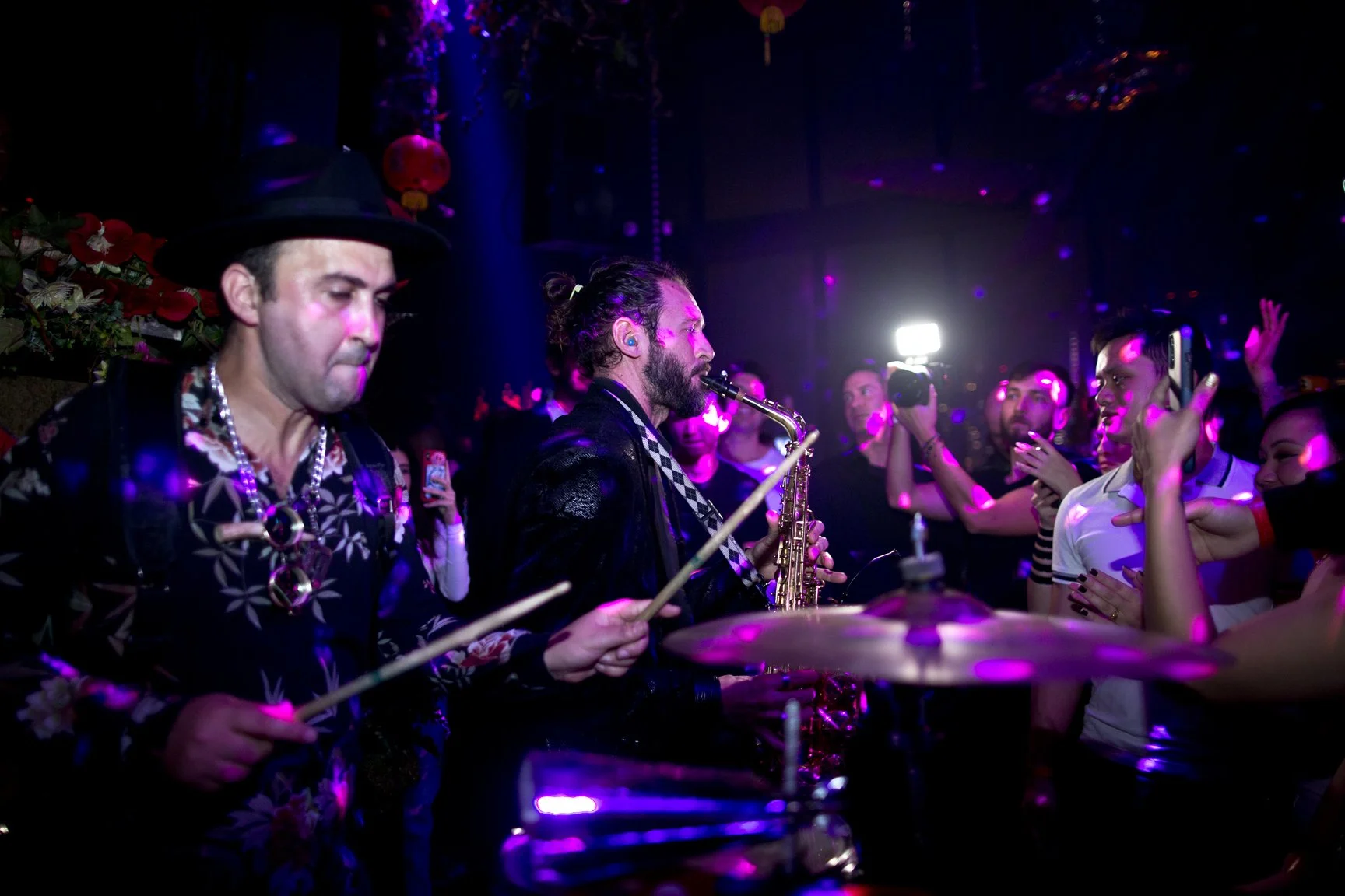 Musicians performing at a lively nightclub with vibrant pink and purple lighting, crowd dancing and recording with smartphones.