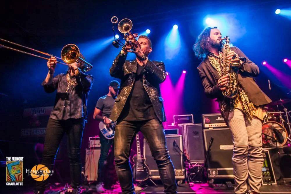 Musicians performing on stage with instruments under colorful stage lighting, including trombone, trumpet, and saxophone, with amplifiers and musical equipment in the background.