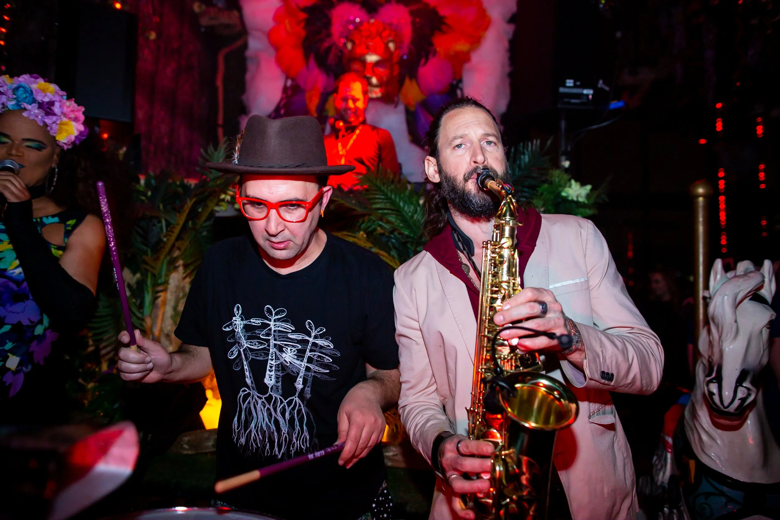 Two musicians performing at a vibrant event with colorful decorations and elaborate costumes, one playing drums and the other playing a saxophone.