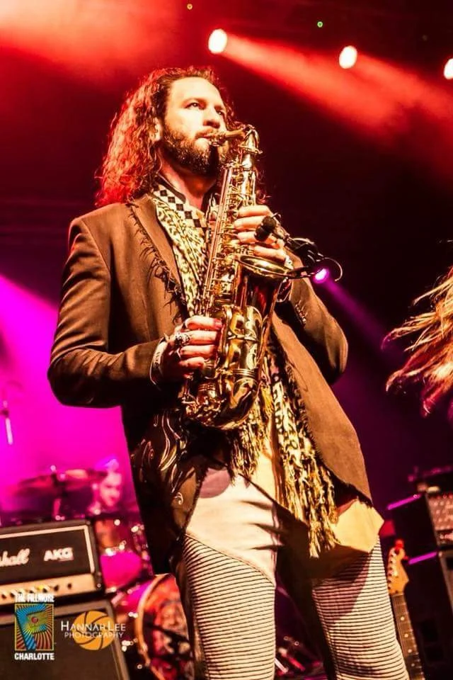 A man with long hair and a beard playing a saxophone on stage with vibrant red, pink, and purple lighting.