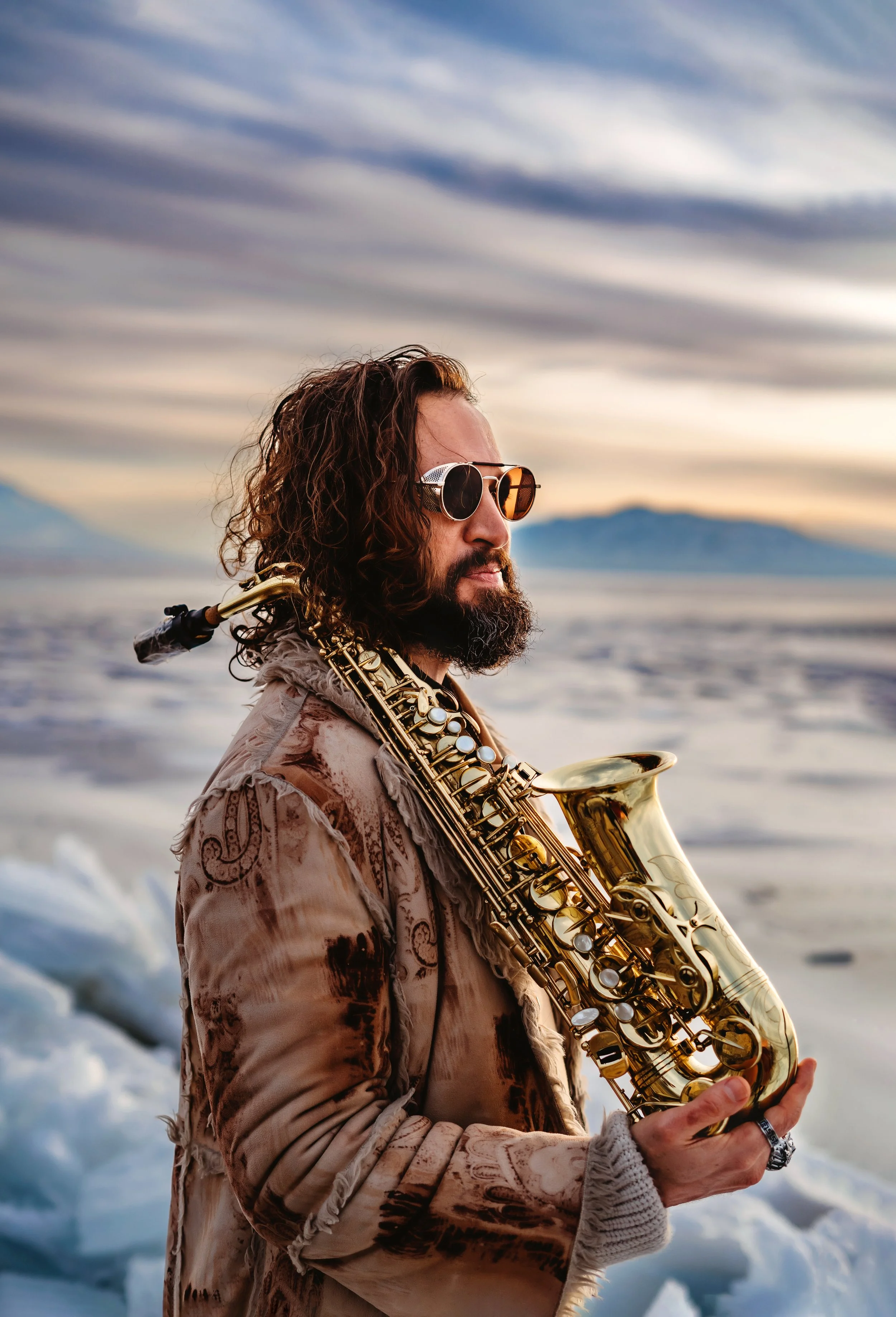 A man with long curly hair, a beard, sunglasses, and a patterned jacket holding a saxophone in a snowy landscape at sunset.