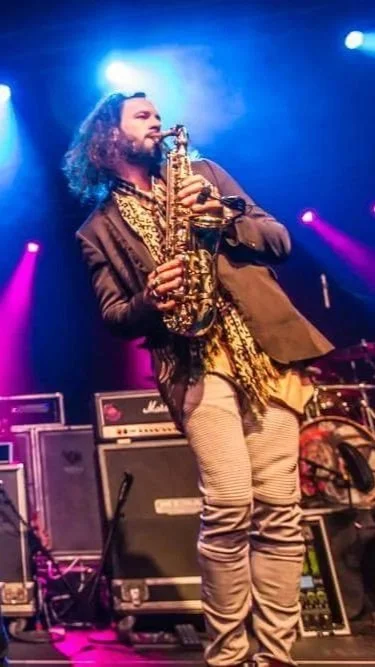 A man with long curly hair and a beard playing a saxophone on stage. The stage is lit with colorful lights, and there are amplifiers and musical equipment around him.