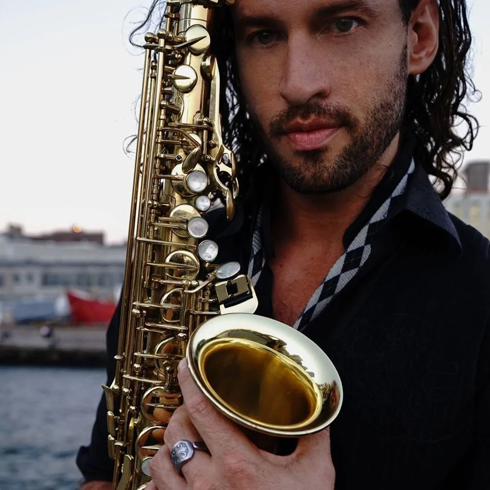 A man with long dark hair and a beard holding a gold saxophone outdoors near a body of water, with boats and buildings in the background.