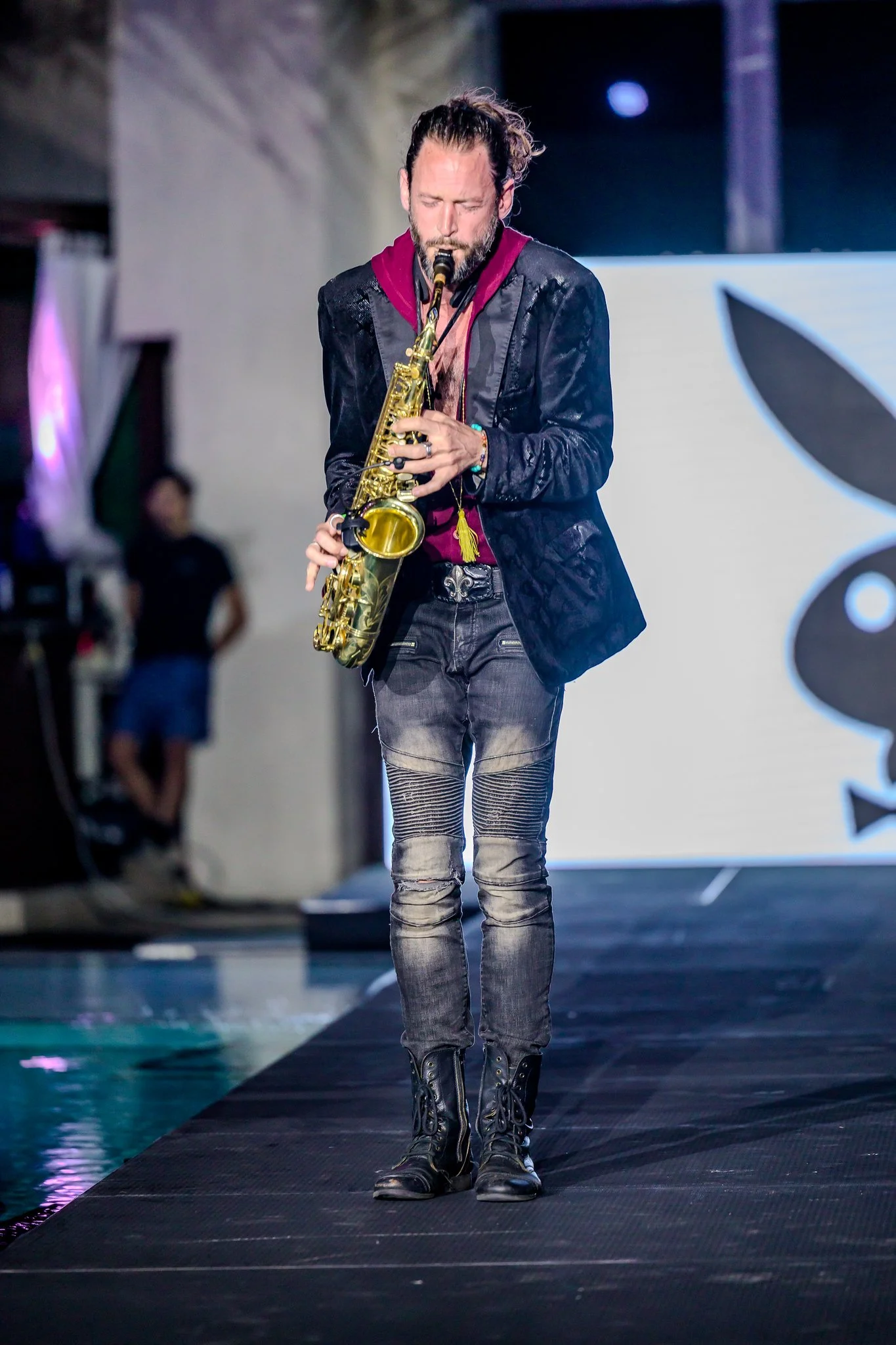 A man with long hair and a beard playing a saxophone on a stage with a white background and a large black and white logo.