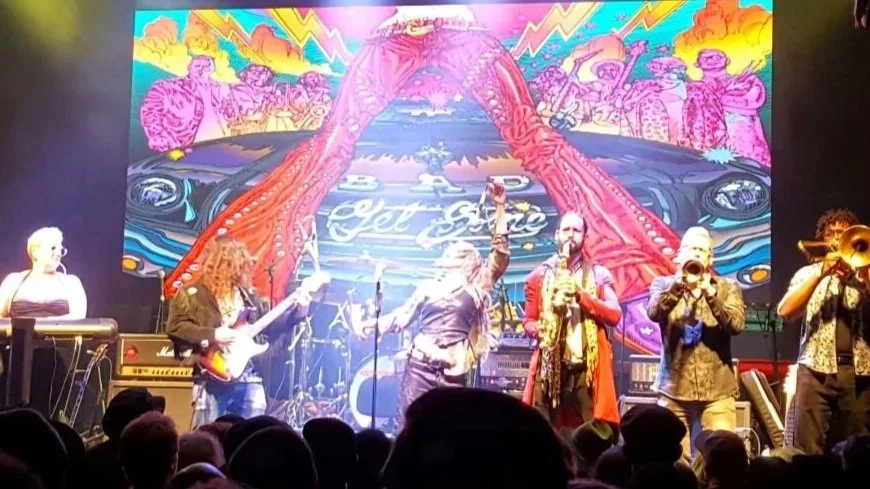 Live band performing on stage with a colorful, psychedelic backdrop and an audience in front.
