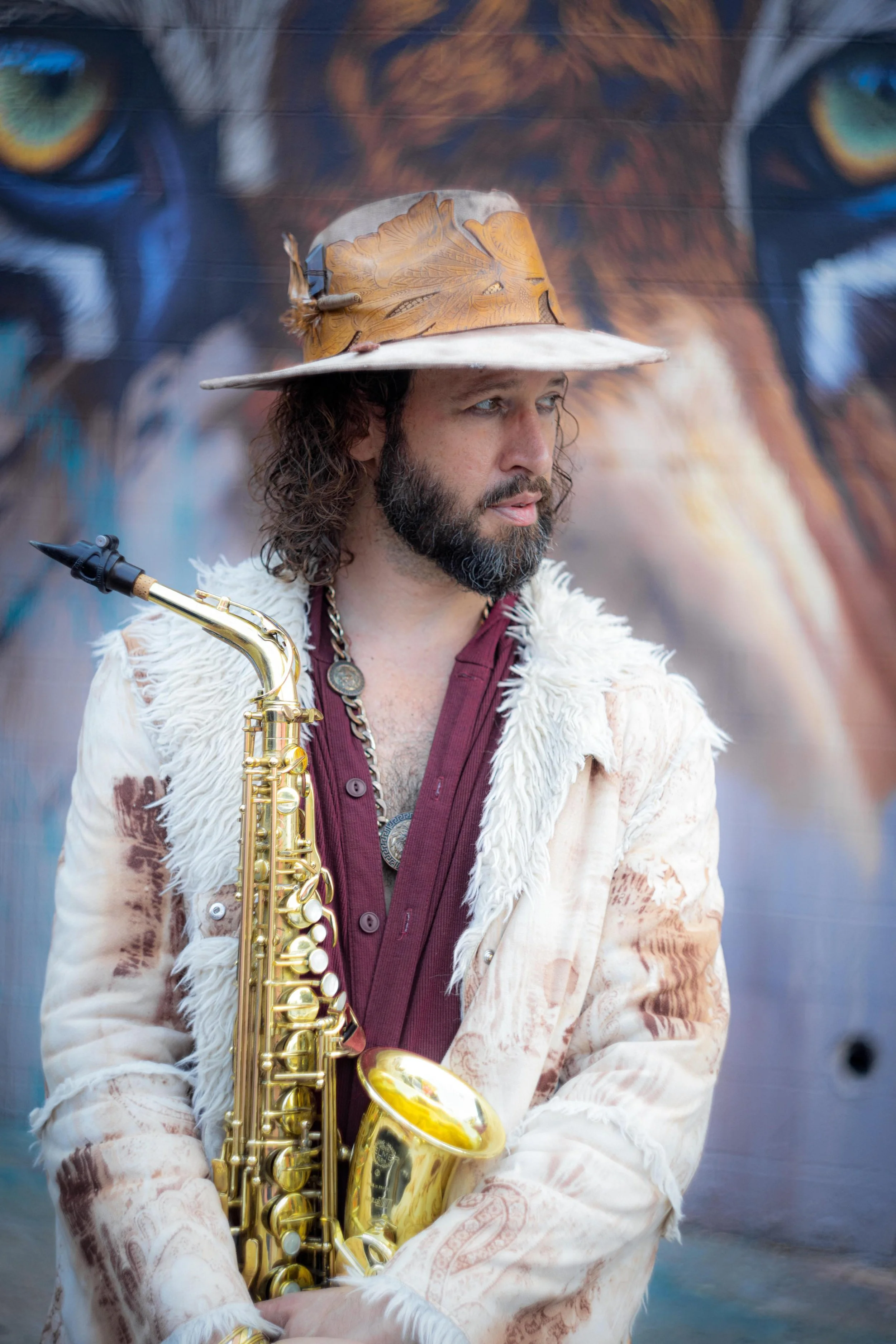 A man with long curly hair and a beard holds a gold saxophone, wearing a beige furry coat, a maroon shirt, and a wide-brimmed hat with feather decorations. Behind him is a large mural of a feline face, possibly a tiger or lion, with striking blue eye