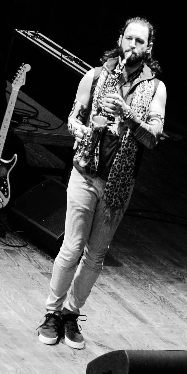 A musician with long hair and beard playing a saxophone on stage, wearing a sleeveless top, patterned scarf, and jeans, with musical equipment and an electric guitar in the background.