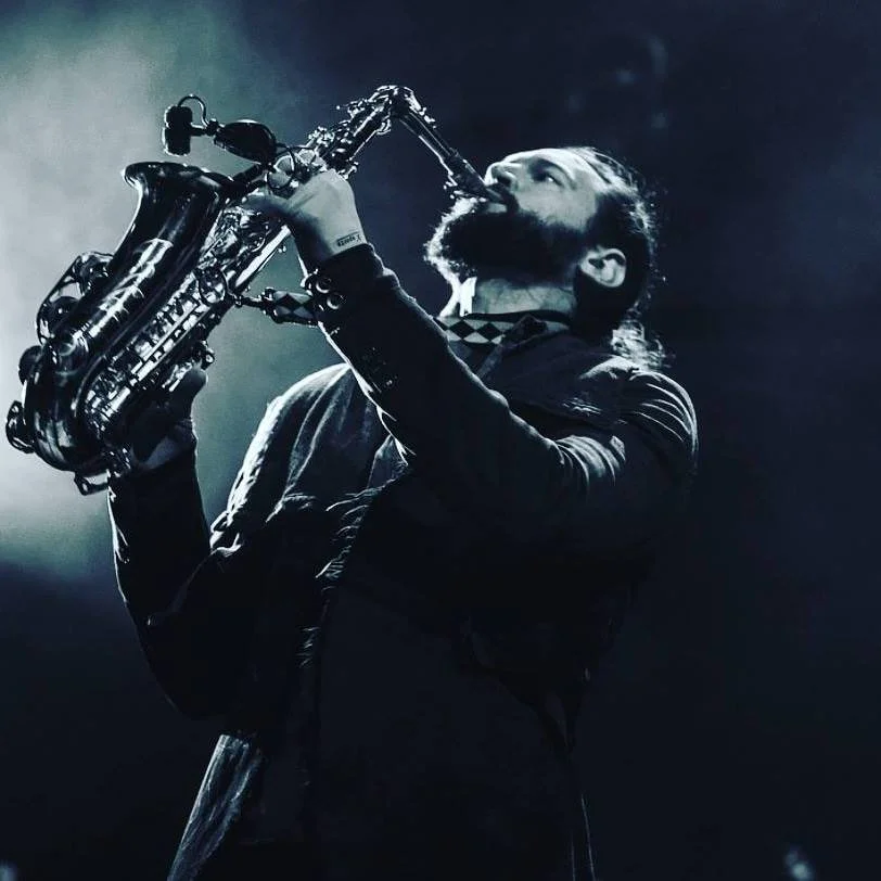 A man with a beard playing a saxophone on stage under dim lighting.